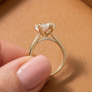 May include: A gold engagement ring with a large, clear gemstone. The ring has a delicate band and is accented with smaller stones. The ring is held against a brown background.