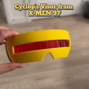 May include: A bright yellow visor with a horizontal red lens, featuring the text Cyclop's Visor from X-MEN 97. The accessory has a smooth, curved frame designed to cover the eye area, with a central cutout for the nose bridge.