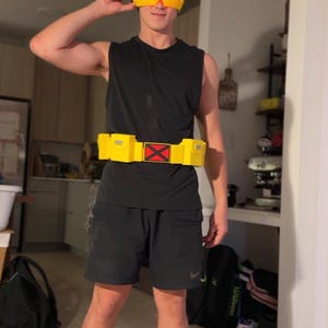 Op de afbeelding: Een geel vizier met een rode lens, een zwart mouwloos shirt, een zwarte korte broek en een gele utility-riem met een centraal rood X-embleem. De riem heeft twee rechthoekige zakjes aan de zijkanten, wat een kostuum van een personage compleet maakt.