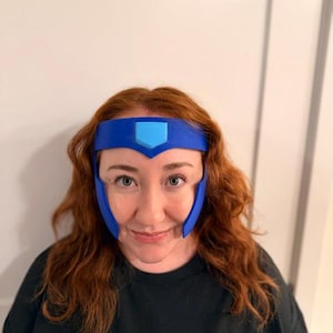 May include: A blue headband featuring a central light blue shield-shaped emblem and two downward-pointing side panels that frame the face. The accessory is constructed from a smooth, solid material and sits across the forehead.