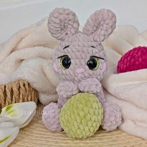 May include: A handmade crochet bunny in soft lavender with large, glittery eyes and a green ball. The bunny is sitting on a white blanket, with a pink ball and a woven basket in the background. The text "SNOWFOXCROCHET" is visible.