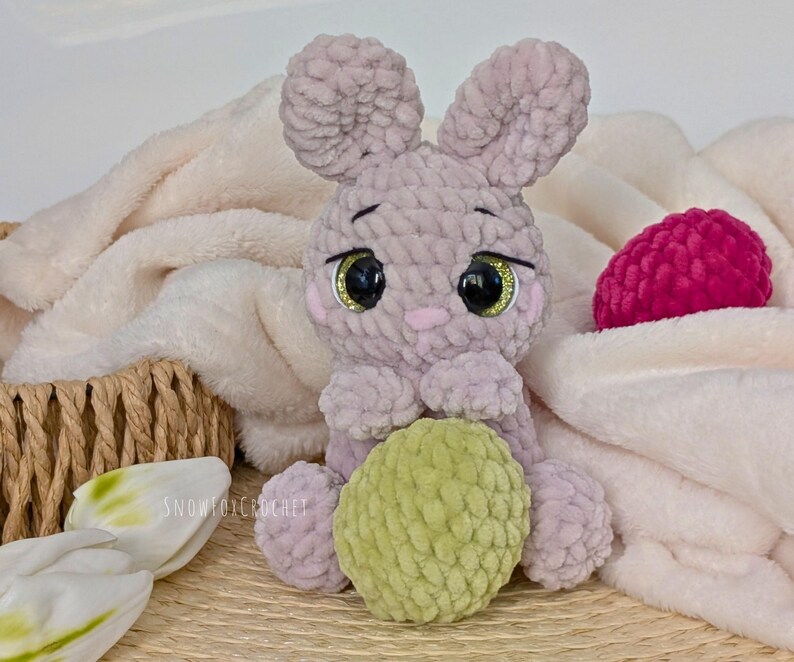 May include: A handmade crochet bunny in soft lavender with large, glittery eyes and a green ball. The bunny is sitting on a white blanket, with a pink ball and a woven basket in the background. The text "SNOWFOXCROCHET" is visible.