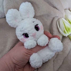 May include: A handmade white crochet bunny with pink accents on the cheeks and around the eyes. The bunny has large, fluffy ears and a long body. The eyes are black with a pink glitter outline. The bunny is a soft, plush toy.