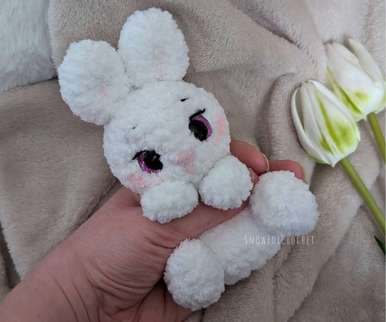 May include: A handmade white crochet bunny with pink accents on the cheeks and around the eyes. The bunny has large, fluffy ears and a long body. The eyes are black with a pink glitter outline. The bunny is a soft, plush toy.