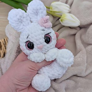 May include: A handmade white crochet bunny with a pink bow and rosy cheeks. The bunny has large black eyes with pink glitter accents. The bunny is soft and plush, perfect for a child's toy or Easter decor.