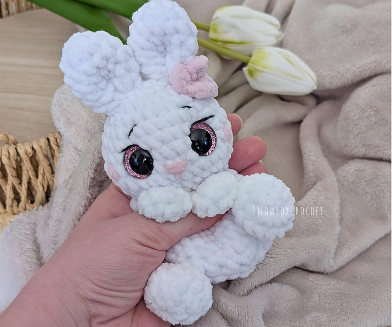 May include: A handmade white crochet bunny with a pink bow and rosy cheeks. The bunny has large black eyes with pink glitter accents. The bunny is soft and plush, perfect for a child's toy or Easter decor.