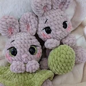 May include: Two handmade crochet bunny plushies. The bunnies are light purple with pink accents on their ears and cheeks. They have large black eyes with gold glitter and are holding a green ball. The background is a soft, white blanket.