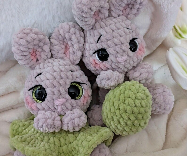 May include: Two handmade crochet bunny plushies. The bunnies are light purple with pink accents on their ears and cheeks. They have large black eyes with gold glitter and are holding a green ball. The background is a soft, white blanket.