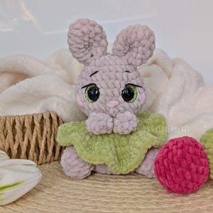 May include: A handmade crochet bunny with large, expressive eyes and a green collar. The bunny is light purple and sits next to a pink and green ball. The background includes a white blanket and a woven basket.