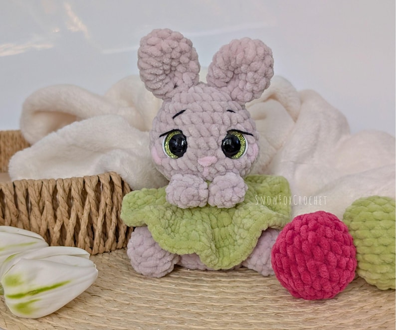 May include: A handmade crochet bunny with large, expressive eyes and a green collar. The bunny is light purple and sits next to a pink and green ball. The background includes a white blanket and a woven basket.