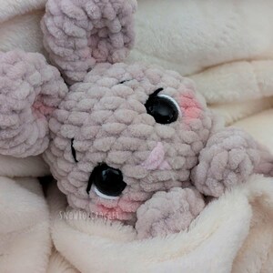 May include: A handmade, light gray crochet bunny toy with large black eyes and pink accents on the cheeks and ears. The bunny is nestled in a soft, white blanket. The text "SNOWFOXCROCHEE" is visible on the blanket.