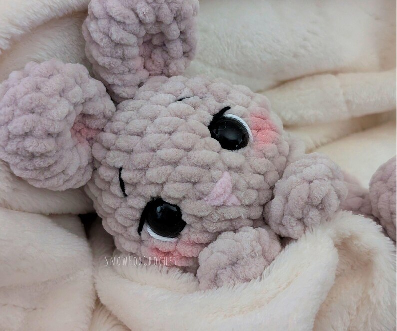 May include: A handmade, light gray crochet bunny toy with large black eyes and pink accents on the cheeks and ears. The bunny is nestled in a soft, white blanket. The text "SNOWFOXCROCHEE" is visible on the blanket.