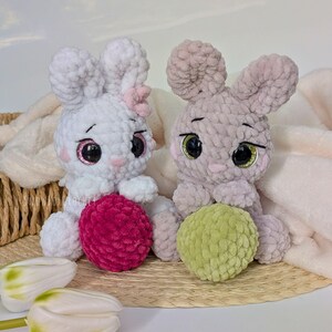 May include: Two handmade crochet bunny plushies. One is white with a pink bow and a magenta ball, the other is gray with a green ball. Both have large, expressive eyes and are sitting on a woven mat.