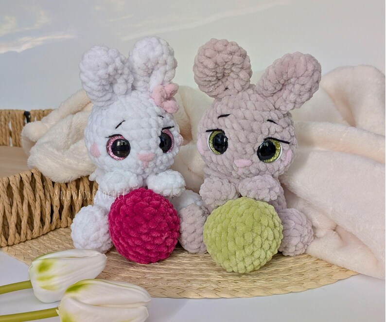 May include: Two handmade crochet bunny plushies. One is white with a pink bow and a magenta ball, the other is gray with a green ball. Both have large, expressive eyes and are sitting on a woven mat.