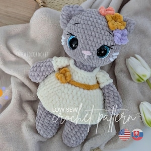 Puede incluir: Muñeca de gato de ganchillo gris hecha a mano con un vestido blanco con cinturón y flor amarillos. El gato tiene ojos azules y una corona de flores. El texto "LOW SEW crochet pattern" es visible. La muñeca está sobre una manta suave y clara.