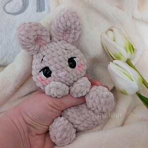 May include: A handmade, light gray crochet bunny with black eyes and pink cheeks. The bunny has large, floppy ears and is holding a soft, plush toy. The background includes white tulips and a fluffy blanket.