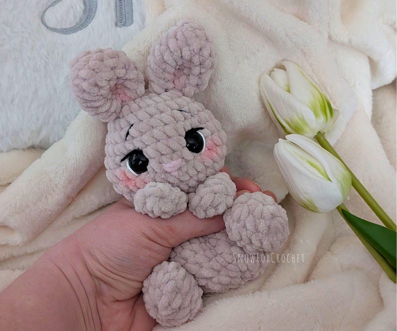 May include: A handmade, light gray crochet bunny with black eyes and pink cheeks. The bunny has large, floppy ears and is holding a soft, plush toy. The background includes white tulips and a fluffy blanket.