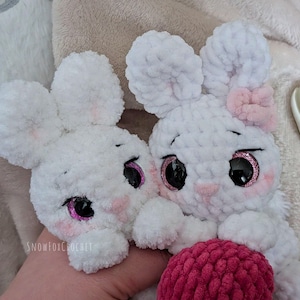 May include: Two white crochet bunny toys with large ears, pink cheeks, and sparkly black eyes. One bunny has a pink bow. The bunnies are holding a pink ball. The text "snowfoxcrochet" is visible on the bottom left.