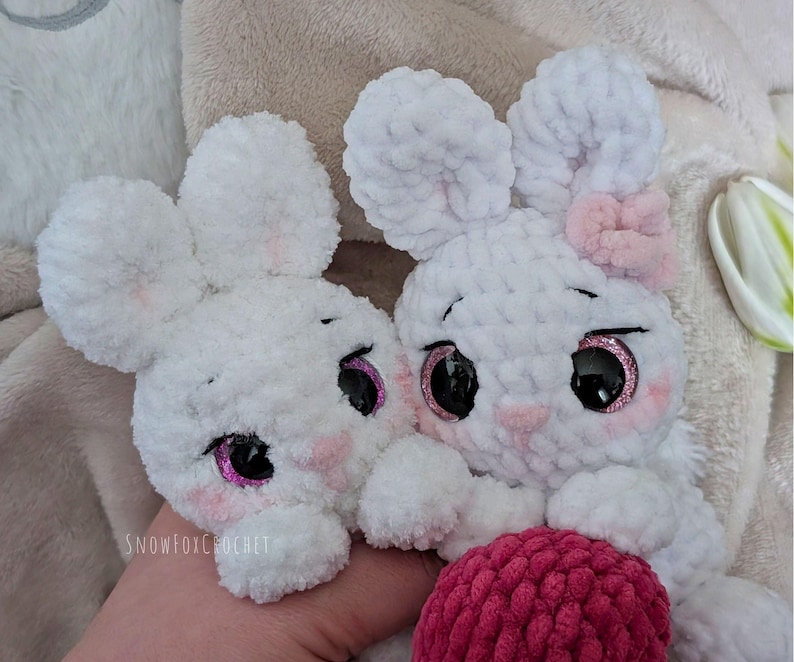 May include: Two white crochet bunny toys with large ears, pink cheeks, and sparkly black eyes. One bunny has a pink bow. The bunnies are holding a pink ball. The text "snowfoxcrochet" is visible on the bottom left.