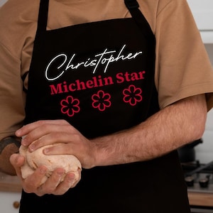 May include: Black apron featuring the name "Christopher", the words "Michelin Star", and three pink flower graphics. The apron is worn by a person in a kitchen setting.