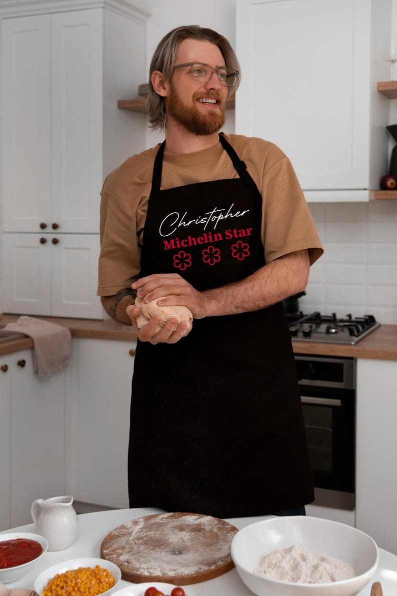 May include: Black apron featuring the name "Christopher", the words "Michelin Star", and three pink flower graphics. The apron is worn by a person in a kitchen setting.