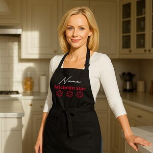 May include: Black apron featuring the text "Name Michelin Star" and three pink flower graphics. The apron has a tie at the waist and two front pockets. The person in the image is wearing a white long-sleeved top.