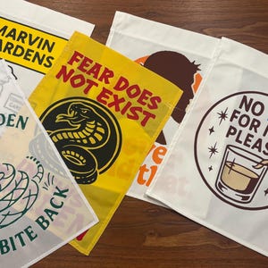 K&ouml;nnte beinhalten: Eine Sammlung dekorativer Gartenflaggen mit verschiedenen Designs und Texten. Eine Flagge zeigt "MARVIN GARDENS". Eine andere zeigt "FEAR DOES NOT EXIST" mit einer Schlangen-Grafik. Weitere Flaggen zeigen S&auml;tze und Illustrationen.
