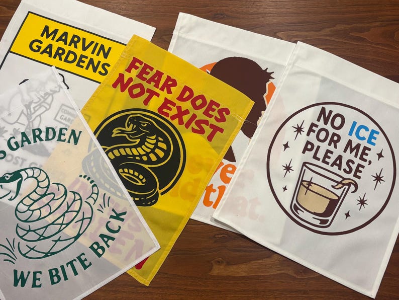 K&ouml;nnte beinhalten: Eine Sammlung dekorativer Gartenflaggen mit verschiedenen Designs und Texten. Eine Flagge zeigt "MARVIN GARDENS". Eine andere zeigt "FEAR DOES NOT EXIST" mit einer Schlangen-Grafik. Weitere Flaggen zeigen S&auml;tze und Illustrationen.