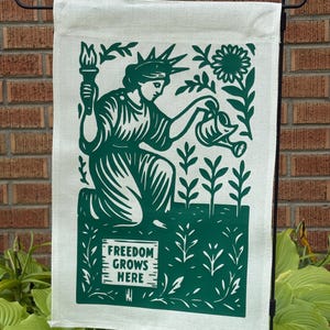 K&ouml;nnte beinhalten: Eine Gartenflagge mit gr&uuml;nem Design auf cremefarbenem Hintergrund. Das Design zeigt die Freiheitsstatue beim Gie&szlig;en von Pflanzen, eine Sonnenblume und den Text "FREEDOM GROWS HERE". Die Flagge h&auml;ngt an einem schwarzen Metallst&auml;nder.