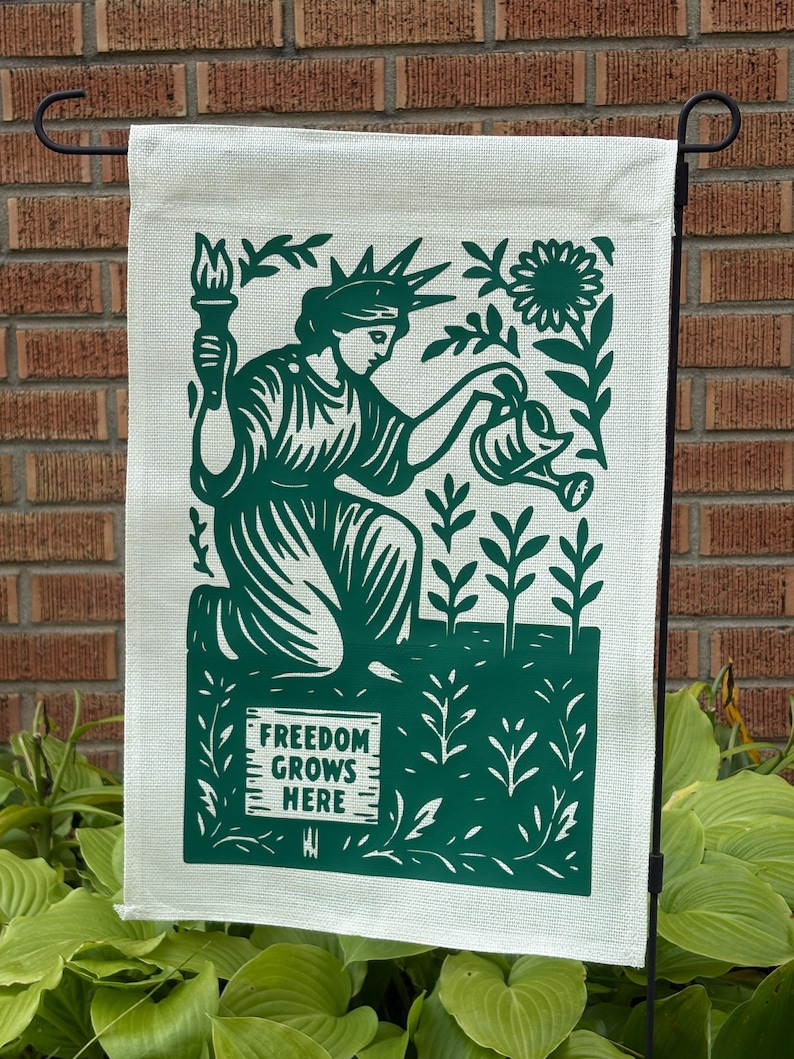 K&ouml;nnte beinhalten: Eine Gartenflagge mit gr&uuml;nem Design auf cremefarbenem Hintergrund. Das Design zeigt die Freiheitsstatue beim Gie&szlig;en von Pflanzen, eine Sonnenblume und den Text "FREEDOM GROWS HERE". Die Flagge h&auml;ngt an einem schwarzen Metallst&auml;nder.