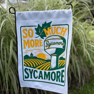 Könnte beinhalten: Weiße Gartenflagge mit grünem und gelbem Design. Die Flagge zeigt die Worte "SO MUCH MORE Sycamore" mit einer Grafik eines Wasserturms, Feldern und einer untergehenden Sonne. Die Flagge hängt an einem schwarzen Metallständer.
