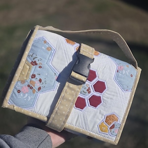 May include: A quilted, handmade bag with a cream-colored background and a beige strap. The bag features a geometric pattern of hexagons in blue, red, and yellow, with floral accents. A black buckle secures the bag's flap.