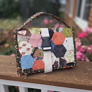 May include: A quilted fabric bag featuring a patchwork of hexagonal patterns in various colors including orange, blue, green, and white with snail and star motifs. The bag has a black handle, a beige webbing strap, and a black plastic side-release buckle.