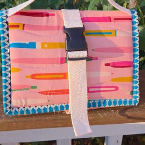 May include: A rectangular quilted fabric case featuring a pink background with a colorful pattern of pens and pencils. The case has a white woven handle and a central white strap secured by a black plastic side-release buckle, with blue trim.