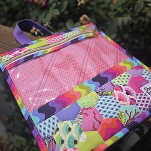 May include: A rectangular zippered pouch featuring a clear vinyl window and a lower section made of multi-colored fabric hexagons with various patterns. The pouch includes a purple fabric handle and a zipper closure along the top edge.