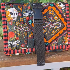 May include: A rectangular quilted fabric bag featuring a dark background with a pattern of white skulls, moths, and orange flowers. The bag has a black plastic buckle closure, a black fabric handle, and red and black striped fabric trim edges.