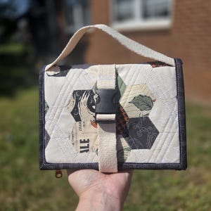 May include: A quilted, rectangular bag with a cream-colored background and hexagonal patches in various patterns and colors. The bag has a dark gray border, a cream-colored handle, and a black buckle closure. The word "ALE" is visible on one of the patches.