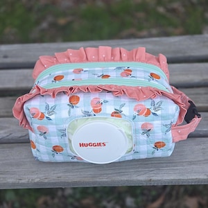 May include: A diaper bag with a light blue and white checkered pattern, adorned with orange fruit and green leaves. The bag features a coral pink ruffle trim and a mint green zipper. A white Huggies wipe container is visible.