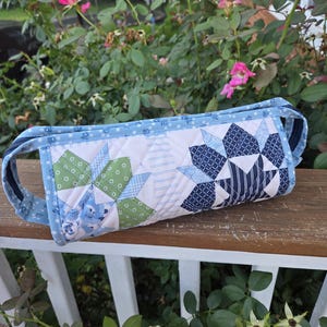May include: A cylindrical quilted fabric case featuring a patchwork design with star-shaped patterns in shades of green, navy blue, and light blue. The case has a light blue floral print trim, a zippered closure, and visible white stitching.