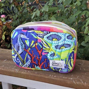 May include: A rectangular quilted zippered pouch featuring a vibrant underwater print with seahorses, coral, and anemones in shades of blue, green, and pink. A white fabric label on the front displays the text Sew RISSA with a small ghost icon.