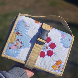 May include: A quilted fabric pouch featuring a white base with hexagonal patches in light blue, red, and orange floral patterns. The pouch has a yellow polka-dot trim, a top handle, and a black plastic side-release buckle closure on the front.