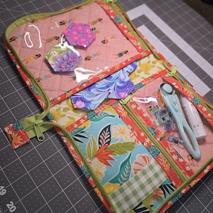 May include: A quilted fabric organizer case featuring a pink background with a hula dancer print, green trim, and multiple zippered clear vinyl pockets. Inside are fabric hexagons, a rotary cutter, sewing clips, and a small white glue stick.