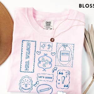 May include: A light pink t-shirt with blue doodles and the text "MRS. WILSON" and "LET'S LEARN". The shirt features illustrations of an open book, lightbulb, pencil, and other school-related icons. A gold necklace is draped across the shirt.