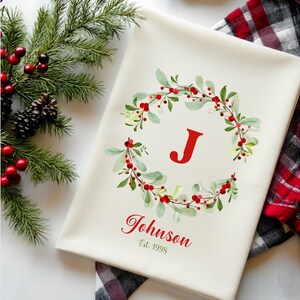 May include: A rectangular off-white kitchen towel featuring a watercolor wreath of green leaves and red berries. In the center is a large red letter J. Below the wreath, the text reads Johnson and Est. 1998 in a red script font.