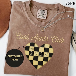 May include: An espresso-coloured t-shirt featuring the text Cool Aunts Club in a light script font. Below the text is a heart-shaped graphic with a black and yellow checkered pattern, the word since, and the year 2026. A black circle with the text CUSTOMIZE YEAR is also present.