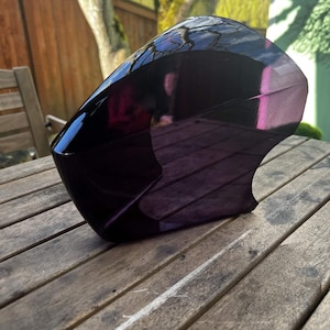 May include: A glossy, dark purple motorcycle fairing, with a curved, aerodynamic design. The fairing is resting on a weathered wooden table, reflecting the surrounding environment. The fairing appears to be made of a durable, lightweight material.