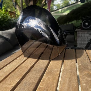 May include: A glossy black, faceted, sculptural object with a smooth, reflective surface. The item features multiple angular planes that catch the light, creating bright white highlights across its dark, curved, and polished exterior form.