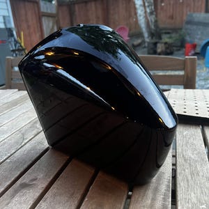May include: A glossy black motorcycle fuel tank with a smooth, curved, and aerodynamic shape. The surface is highly reflective, showing highlights from the surrounding environment across its dark, polished finish and contoured exterior design.