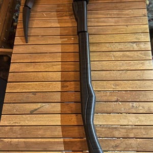 Può includere: Una falce nera con una lama curva e un lungo manico, appoggiata su un tavolo di legno. La falce sembra essere fatta di un materiale scuro, forse metallico, ed è progettata con angoli acuti e parti segmentate.