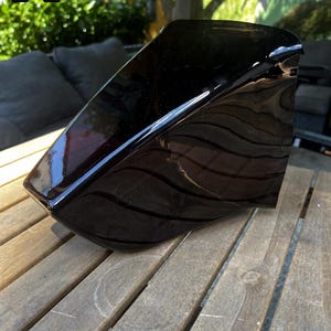 May include: A glossy black, angular, molded plastic component with a smooth, reflective surface. The object features a distinct, sharp-edged geometric shape with a curved top profile and a flat base, showcasing a solid, dark, uniform finish.